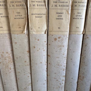 Puede incluir: Una colecci&oacute;n de libros antiguos con cubiertas blanquecinas y etiquetas de papel envejecido. Los t&iacute;tulos incluyen "The Little Minister", "Sentimental Tommy" y "Tommy and Grizel". Las lomos muestran signos de desgaste y manchas.