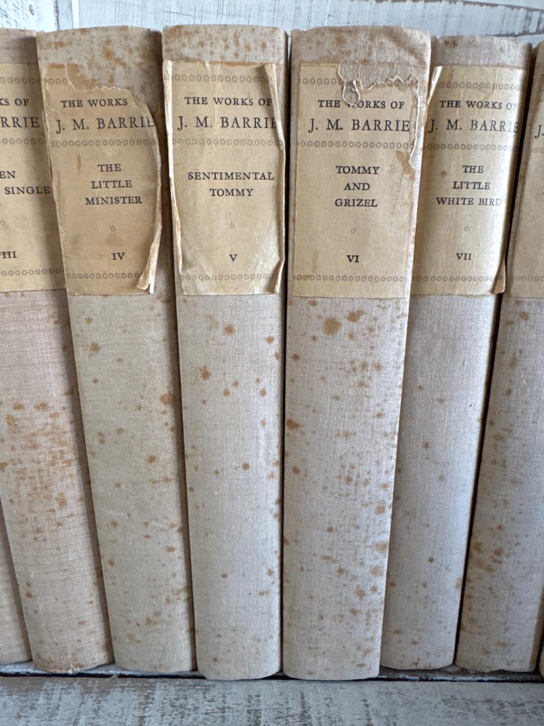 Puede incluir: Una colecci&oacute;n de libros antiguos con cubiertas blanquecinas y etiquetas de papel envejecido. Los t&iacute;tulos incluyen "The Little Minister", "Sentimental Tommy" y "Tommy and Grizel". Las lomos muestran signos de desgaste y manchas.