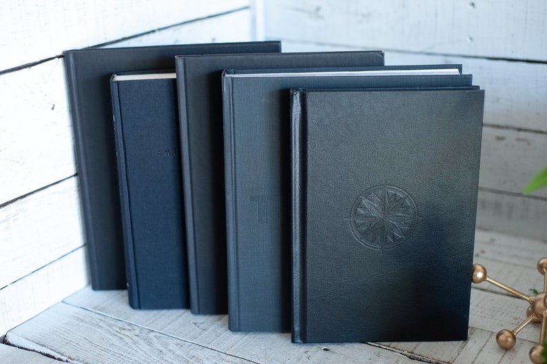 May include: A collection of five dark-colored books, varying in size and thickness, are arranged in a row. The books have textured covers, with one featuring a compass rose design. The spines are visible, and some have subtle embossed details.