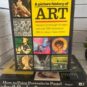 K&ouml;nnte beinhalten: Ein schwarzes Hardcover-Buch mit dem Titel "A picture history of ART" mit gelbem Hintergrund. Der Buchumschlag zeigt eine Vielzahl von bunten Illustrationen von Kunst aus verschiedenen Epochen. Das Buch liegt auf einem anderen Buch mit dem Titel "How to Paint Portraits in Pastel" von Norman Rockwell.