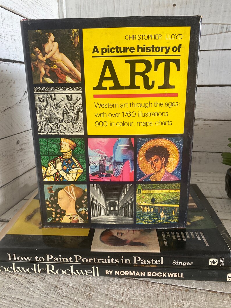 K&ouml;nnte beinhalten: Ein schwarzes Hardcover-Buch mit dem Titel "A picture history of ART" mit gelbem Hintergrund. Der Buchumschlag zeigt eine Vielzahl von bunten Illustrationen von Kunst aus verschiedenen Epochen. Das Buch liegt auf einem anderen Buch mit dem Titel "How to Paint Portraits in Pastel" von Norman Rockwell.