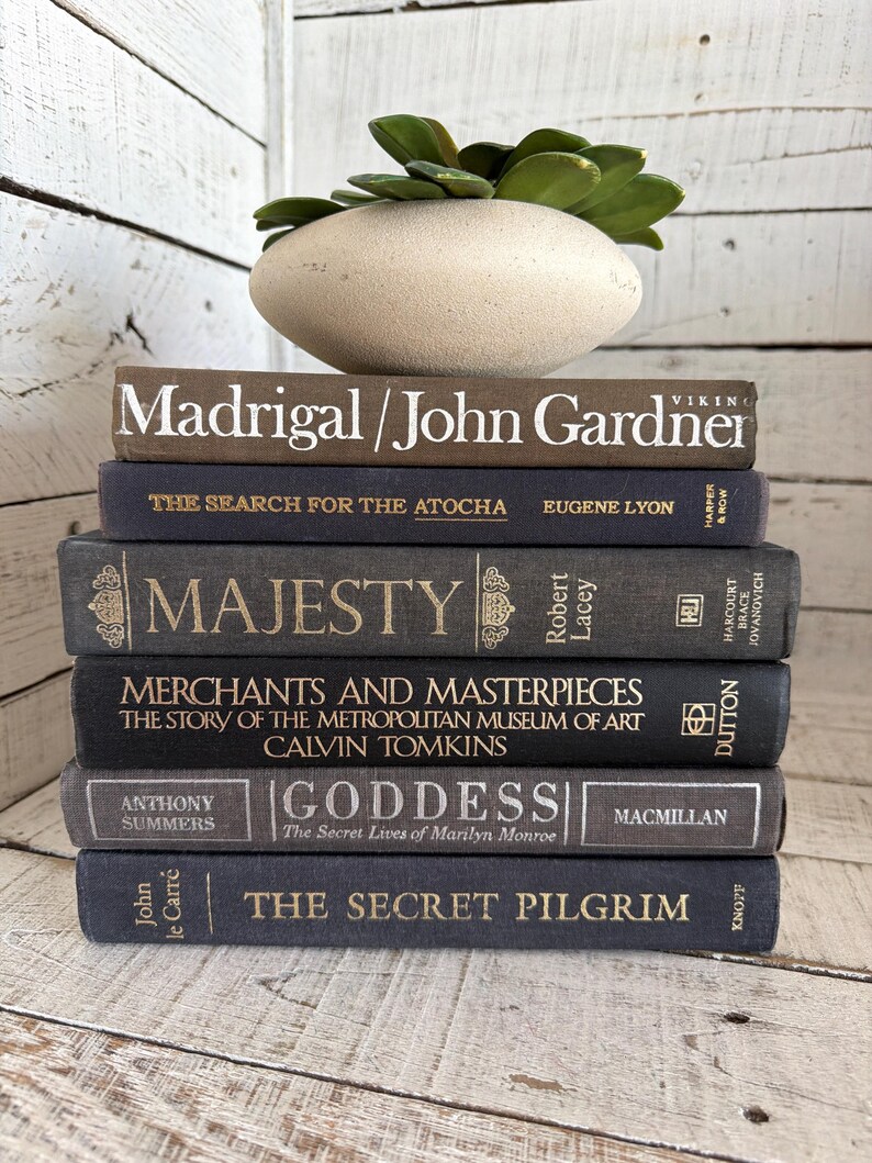 May include: A collection of six hardback books in shades of grey and navy blue, with gold lettering. Titles include "Madrigal/John Gardner" and "The Secret Pilgrim." A decorative, oval-shaped, beige stone pot with green plant sits atop the books.