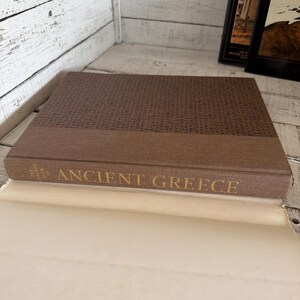 May include: A brown hardcover book titled The Horizon Book of Ancient Greece with a textured fabric cover. The front cover features a repeating pattern of Greek lettering in a darker brown shade, while the spine displays the title in gold text.