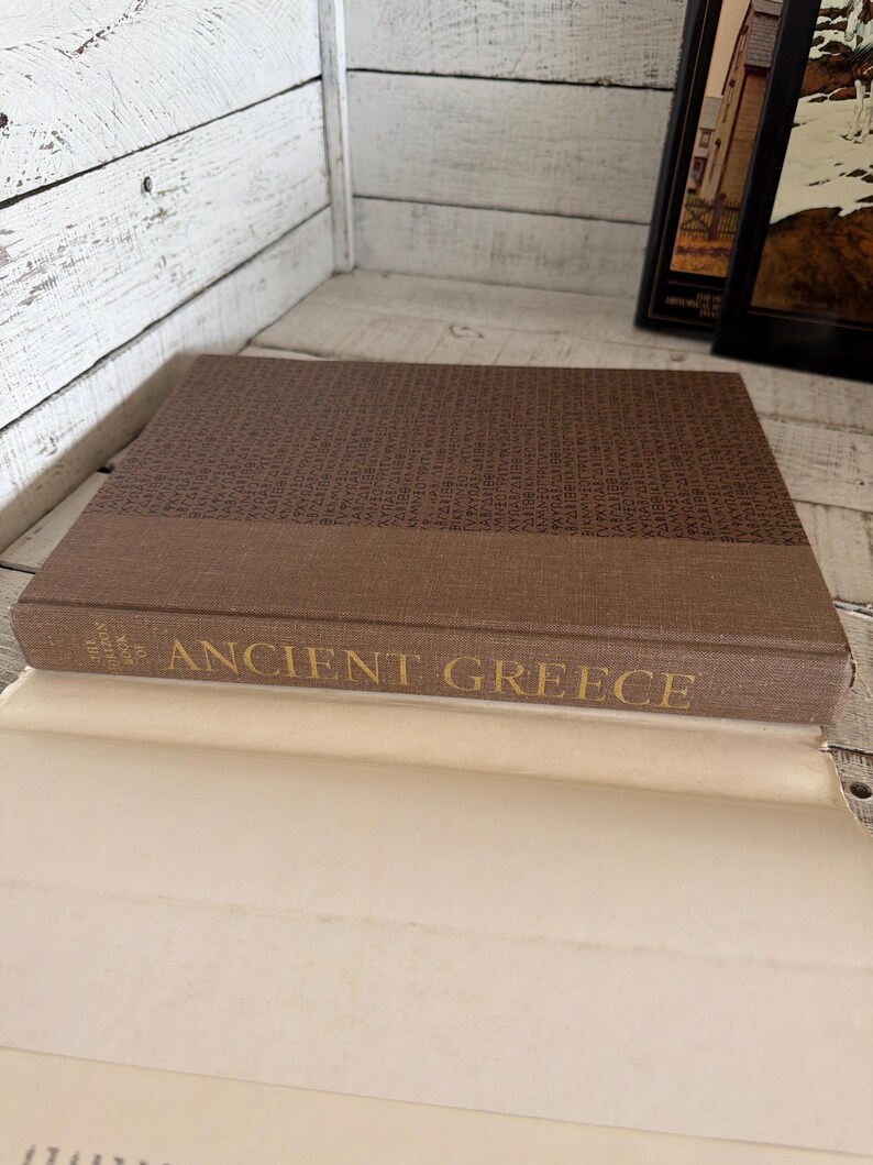 May include: A brown hardcover book titled The Horizon Book of Ancient Greece with a textured fabric cover. The front cover features a repeating pattern of Greek lettering in a darker brown shade, while the spine displays the title in gold text.