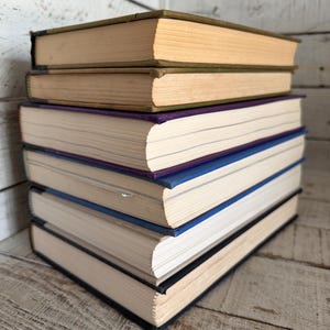 May include: A stack of six hardcover books. The top two books have tan covers and aged pages. The next book has a purple cover, followed by a blue one. The bottom two books have black covers. The books are stacked on a weathered wooden surface.