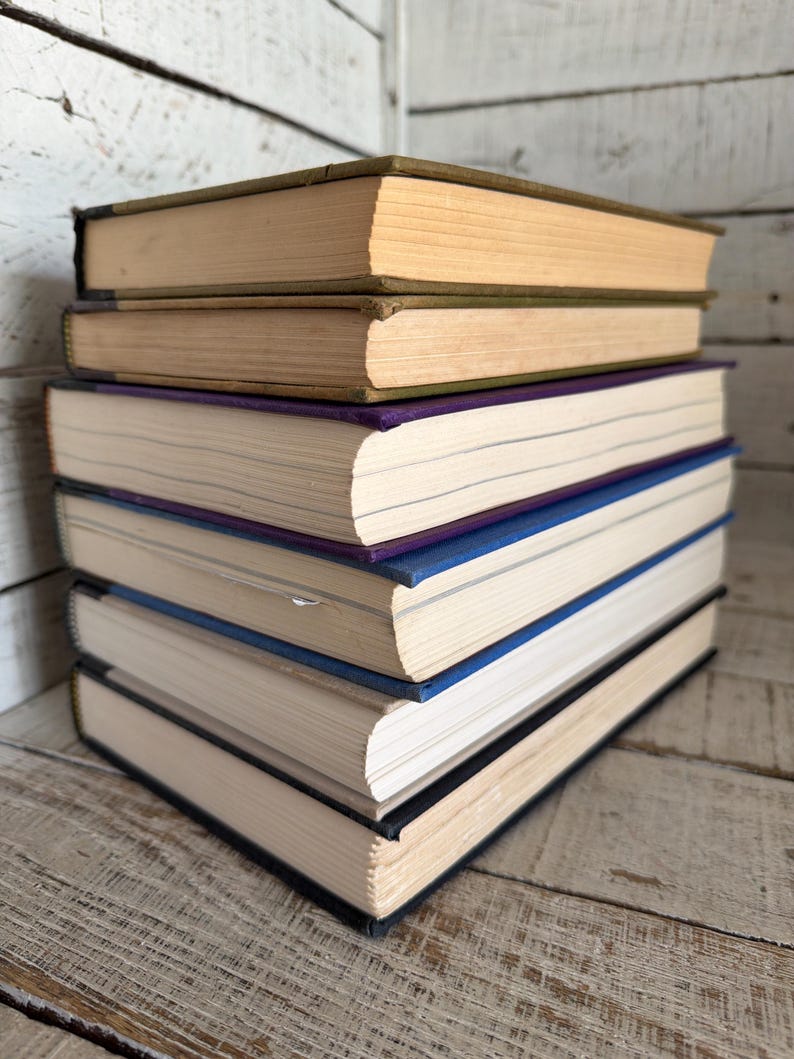 May include: A stack of six hardcover books. The top two books have tan covers and aged pages. The next book has a purple cover, followed by a blue one. The bottom two books have black covers. The books are stacked on a weathered wooden surface.