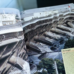 May include: An open book displays a photograph of a collapsed building in Kobe, Japan. The image shows the aftermath of an earthquake, with the structure severely damaged. The text "Atlas Shrugged" is visible.