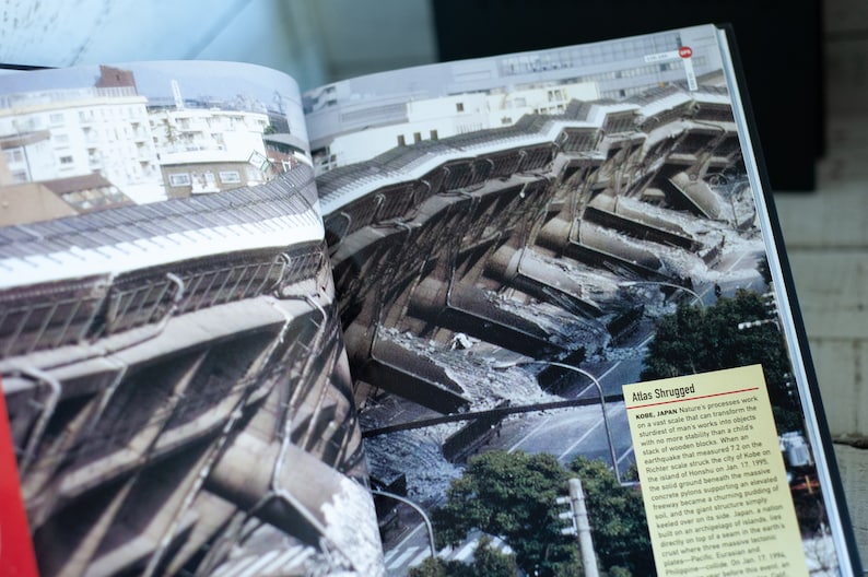 May include: An open book displays a photograph of a collapsed building in Kobe, Japan. The image shows the aftermath of an earthquake, with the structure severely damaged. The text "Atlas Shrugged" is visible.
