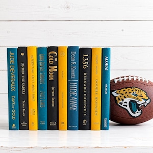 May include: A row of ten hardcover books with spines in shades of teal, black, and yellow, featuring various titles and authors, positioned next to a brown leather American football with a Jacksonville Jaguars logo graphic on the side.