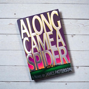 May include: A hardcover book titled "ALONG CAME A SPIDER" by James Patterson. The book cover is purple with silver and yellow lettering. The bottom of the cover features a green field, a house, and a sunset.