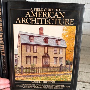 May include: A hardcover book titled A Field Guide to American Architecture by Carole Rifkind. The black cover features a photograph of a historic house with yellow siding, a gray roof, and a chimney, surrounded by autumn trees and a wooden fence.