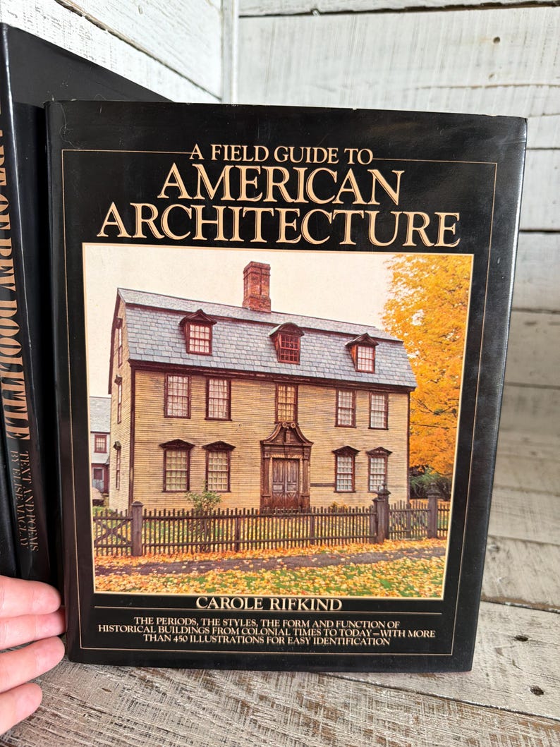May include: A hardcover book titled A Field Guide to American Architecture by Carole Rifkind. The black cover features a photograph of a historic house with yellow siding, a gray roof, and a chimney, surrounded by autumn trees and a wooden fence.