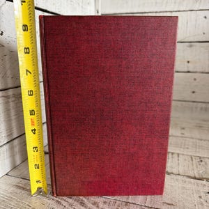 Puede incluir: Un libro de tapa dura de color rojo oscuro con una cubierta texturizada. El libro mide aproximadamente 20 cm de alto, medido con una cinta m&eacute;trica amarilla. El lomo del libro es visible y las p&aacute;ginas no son visibles.