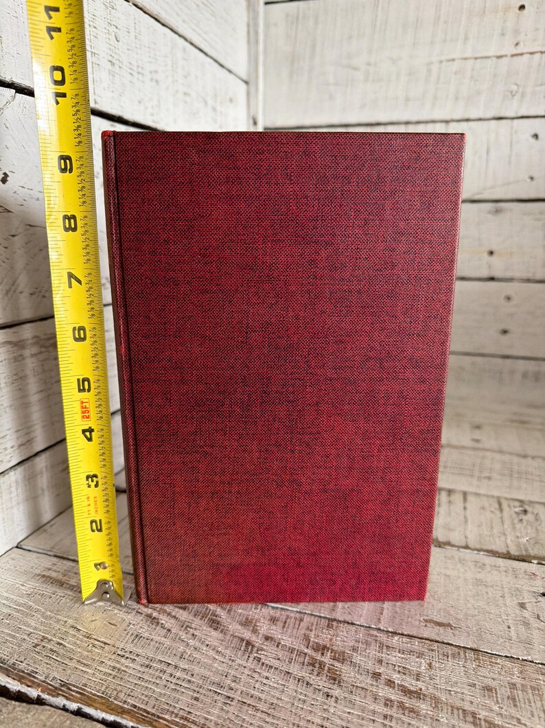 Puede incluir: Un libro de tapa dura de color rojo oscuro con una cubierta texturizada. El libro mide aproximadamente 20 cm de alto, medido con una cinta m&eacute;trica amarilla. El lomo del libro es visible y las p&aacute;ginas no son visibles.