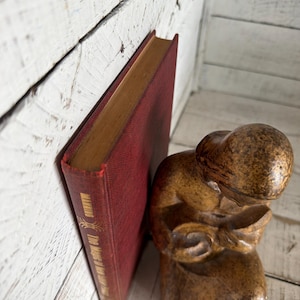 Puede incluir: Un libro rojo vintage titulado "The Meaning of Love" se apoya contra una superficie de madera blanca. Una peque&ntilde;a figura marr&oacute;n tallada de una persona leyendo est&aacute; cerca. El libro tiene letras doradas.