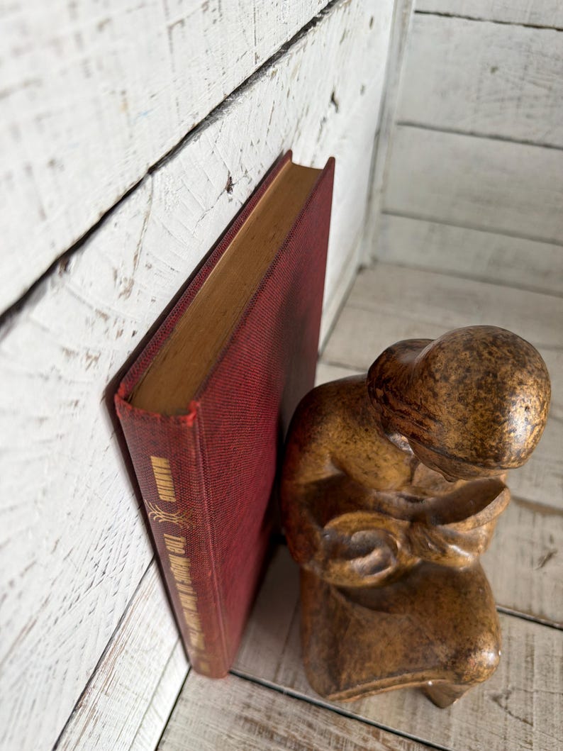 Puede incluir: Un libro rojo vintage titulado "The Meaning of Love" se apoya contra una superficie de madera blanca. Una peque&ntilde;a figura marr&oacute;n tallada de una persona leyendo est&aacute; cerca. El libro tiene letras doradas.