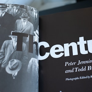 May include: A black and white photograph of a book spread open. The left page features a crowd of men in suits and hats. The right page has the title "The Century" in large white letters, with author names below.