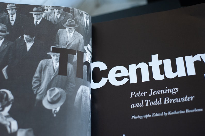 May include: A black and white photograph of a book spread open. The left page features a crowd of men in suits and hats. The right page has the title "The Century" in large white letters, with author names below.