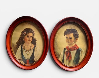 May include: Two oval-shaped wall portraits in dark red wooden frames. The left portrait depicts a person with long dark hair wearing a white blouse, blue vest, and beaded necklace. The right portrait shows a person with short hair wearing a white shirt, blue vest, and a red neckerchief.