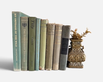 May include: A collection of eight vintage books with muted green, gray, and beige spines arranged in a descending height order, accompanied by a small, ornate, metallic gold-toned vase containing a sprig of dried foliage on the right side.