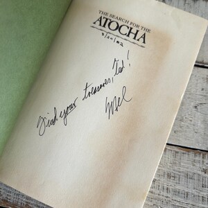 May include: An open book titled "The Search for the Atocha" with a handwritten message. The book has aged pages and a green cover. The message reads "Find your treasures, Ted! Mel".