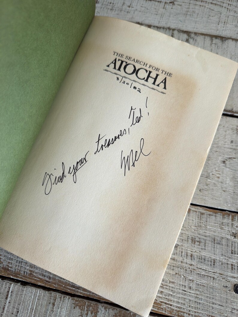 May include: An open book titled "The Search for the Atocha" with a handwritten message. The book has aged pages and a green cover. The message reads "Find your treasures, Ted! Mel".