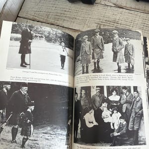 May include: An open book featuring historical black and white photographs. The images depict George V and his son, Prince John, and other members of the royal family in Scotland, wearing kilts and formal clothing.