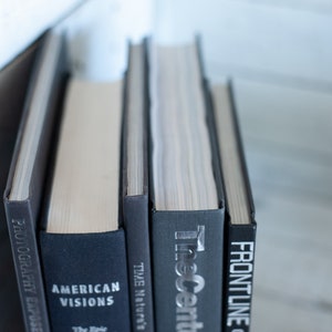 May include: A stack of hardcover books with dark gray and black covers. Titles include "Photography Exposed", "American Visions", "Time Nature's", "The Ascent", and "Frontline". The books are arranged vertically, with the spines facing the viewer.