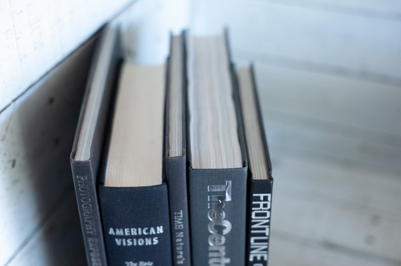 May include: A stack of hardcover books with dark gray and black covers. Titles include "Photography Exposed", "American Visions", "Time Nature's", "The Ascent", and "Frontline". The books are arranged vertically, with the spines facing the viewer.