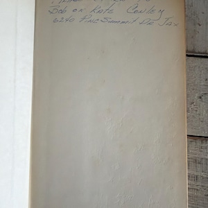 Puede incluir: Libro abierto con texto manuscrito en tinta azul. El texto dice "Please Return to - Bob or Kate Conley 6240 Pine Summit Dr. Jax". Las p&aacute;ginas son blanquecinas con una apariencia texturizada.