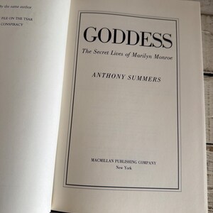 May include: An open book displaying the title "GODDESS" in bold black font. The subtitle reads "The Secret Lives of Marilyn Monroe" by Anthony Summers. The Macmillan Publishing Company is also listed.