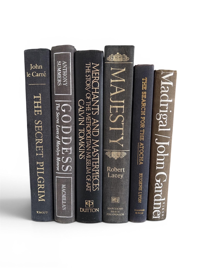 May include: A selection of seven hardback books with spines in shades of grey and brown. Titles include "The Secret Pilgrim," "Goddess," and "Majesty," with gold lettering. The books are arranged vertically against a white backdrop.