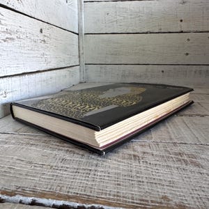 May include: A black hardback book with a gold and black patterned cover. The book is lying on a white wooden surface.