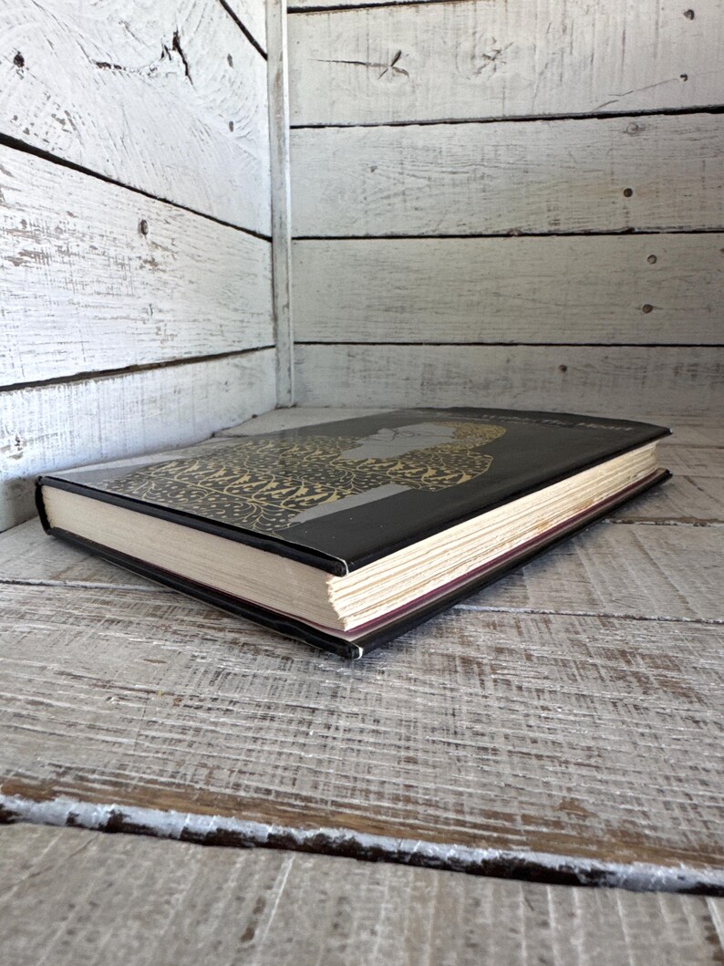 May include: A black hardback book with a gold and black patterned cover. The book is lying on a white wooden surface.