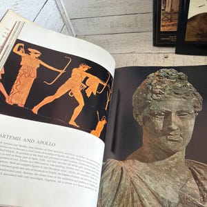 May include: An open book featuring a page with the text ARTEMIS AND APOLLO above an orange and black illustration of two figures with bows. The opposite page displays a photograph of a weathered, green-toned bronze bust of a person.