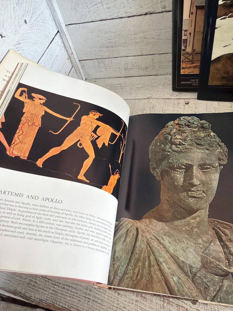 May include: An open book featuring a page with the text ARTEMIS AND APOLLO above an orange and black illustration of two figures with bows. The opposite page displays a photograph of a weathered, green-toned bronze bust of a person.