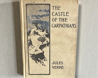 May include: Vintage book titled "The Castle of the Carpathians" by Jules Verne. The cover features a black and white illustration of a castle on a mountain with a blue sky. The book has a beige cover.