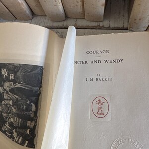 Puede incluir: Libro abierto titulado "Courage Peter and Wendy" de J.M. Barrie. El libro tiene una cubierta blanquecina con texto negro. Se ve un emblema rojo y blanco. Publicado por Charles Scribner's Sons, Nueva York, MCMXXX.