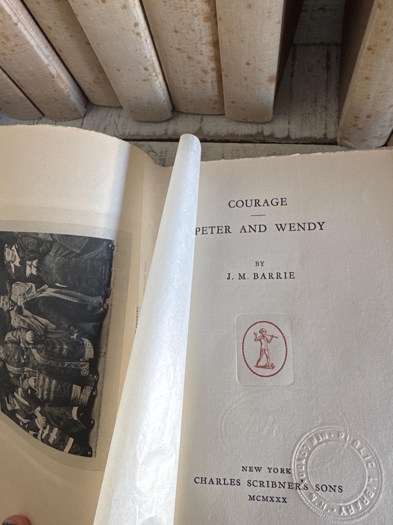 Puede incluir: Libro abierto titulado "Courage Peter and Wendy" de J.M. Barrie. El libro tiene una cubierta blanquecina con texto negro. Se ve un emblema rojo y blanco. Publicado por Charles Scribner's Sons, Nueva York, MCMXXX.