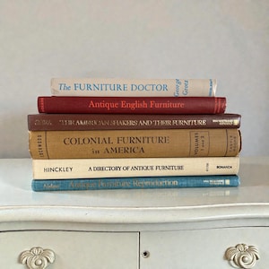 May include: A stack of antique furniture books with titles including "The Furniture Doctor," "Antique English Furniture," and "Colonial Furniture in America." The books are in various colors, including red, brown, and beige, and are stacked on a white surface.