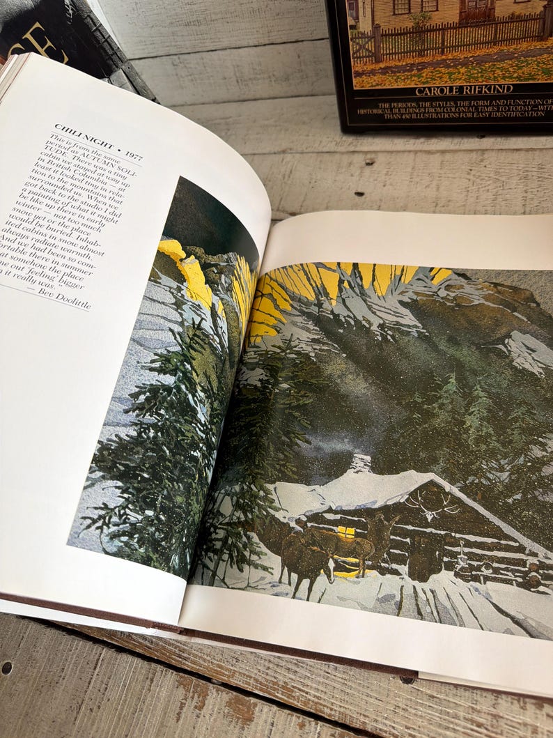 May include: An open book displaying a print titled CHILI NIGHT 1977 by Bev Doolittle. The illustration features a snow-covered cabin at night with dark green pine trees, a bright yellow sky, and two horses standing near the cabin entrance.