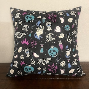 May include: A black throw pillow with a white and blue print of various Disney villain-themed icons, including a poison apple, a crown, and a potion bottle.