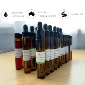 May include: A collection of small brown glass bottles with dropper tops, each containing a different essential oil. The bottles are labelled with the name of the oil, such as "Wintergreen", "Frankincense", "Clove Bud", "Rosemary", "Tea Tree", "Vanilla", "Rose", "Cinnamon", and "Lavender". The bottles are arranged in a row on a wooden surface. The image also includes three icons: a water drop, a map of Australia, and a rabbit. The text "100% Pure Essential Oil", "Australian Made & Owned", and "Cruelty Free" is also visible.
