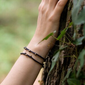 Op de afbeelding: Twee zwarte touwarmbanden met kleine gouden kralen worden om de pols van een persoon gedragen. De hand van de persoon rust op een boomstam. De achtergrond is wazig, met groen gebladerte en natuurlijk licht.