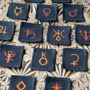 Op de afbeelding: Een verzameling kleine, vierkante, zwarte stoffen patches met oranje astrologische symbolen. De patches zijn gerangschikt op een gebeeldhouwd houten oppervlak, met verschillende hemelse en planetaire symbolen.