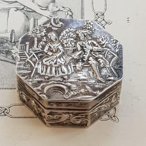 May include: An octagonal silver-toned metal box featuring an embossed scene on the lid of two figures seated on a bench in a garden setting. The sides of the box are decorated with intricate scrollwork and floral relief patterns in a metallic finish.