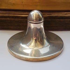 Pode incluir: Um mata-borrão de metal prateado polido com uma cúpula lisa em forma de sino e uma base circular larga. A superfície reflexiva mostra o ambiente distorcido e a base apresenta pequenas marcas de contraste gravadas na borda inferior.