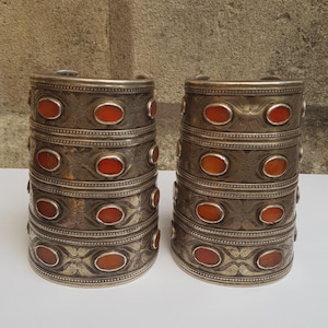 May include: A pair of wide, silver-toned metal cuff bracelets featuring a stacked, multi-band design. Each cuff is decorated with rows of oval-shaped, reddish-orange cabochon stones set within engraved, swirling patterns and beaded metal borders.