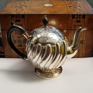 May include: A silver teapot featuring a rounded body with vertical fluted detailing, a curved black handle, and a matching black finial on the lid. The teapot rests on a circular pedestal base and includes a curved spout and engraved hallmarks.