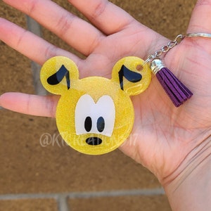 May include: Yellow glitter keychain in the shape of a cartoon character's face, with black ears and a surprised expression. The keychain has a silver chain and a purple tassel. The character's face has white eyes and a black mouth.